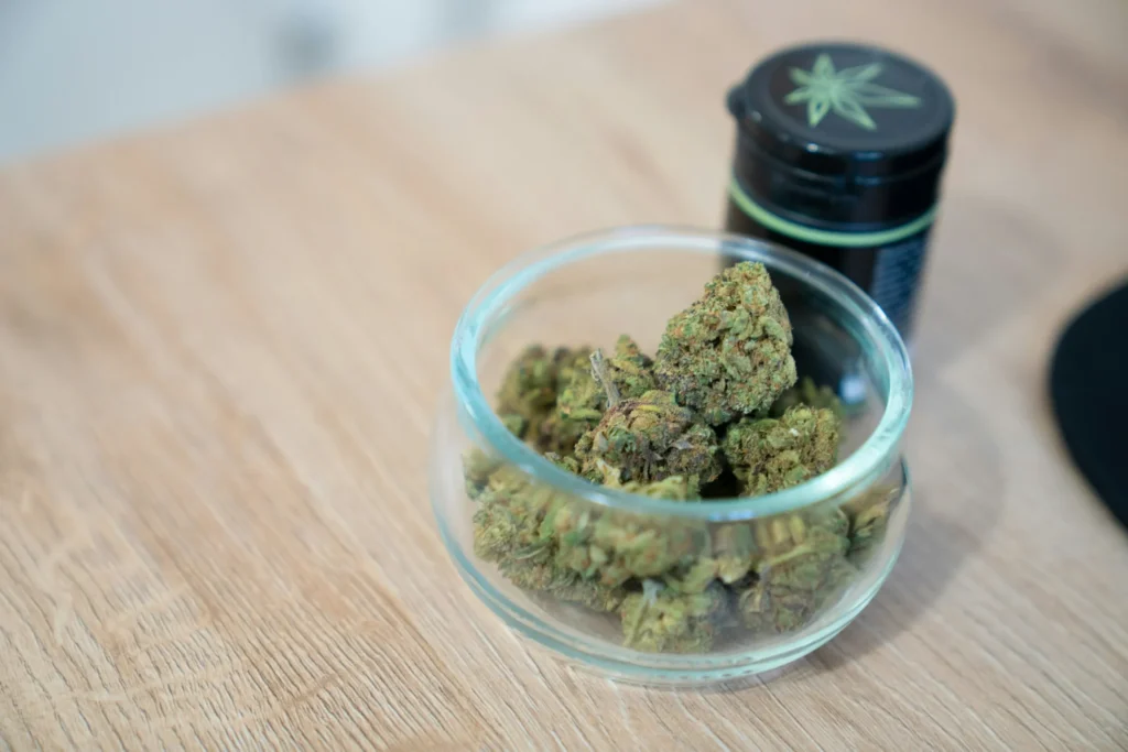 Glass bowl with dried cannabis buds and a black container with a cannabis leaf logo on a wooden table.