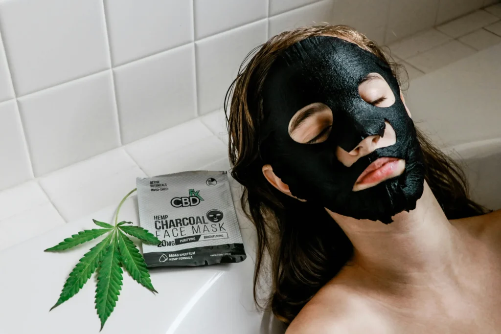 A person relaxes in a bath with a black CBD charcoal face mask, next to a package labeled "Hemp Charcoal Face Mask" and a cannabis leaf.