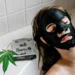 A person relaxes in a bath with a black CBD charcoal face mask, next to a package labeled "Hemp Charcoal Face Mask" and a cannabis leaf.