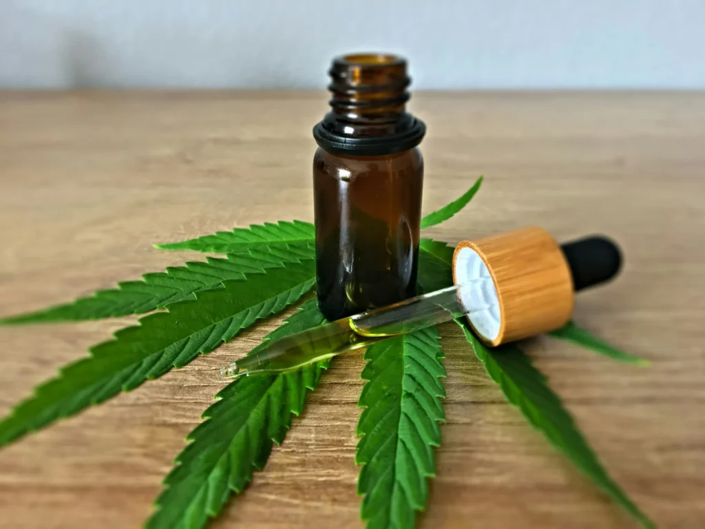 A brown glass dropper bottle of cannabis tincture sits on a wooden surface with green cannabis leaves, its dropper placed beside it.