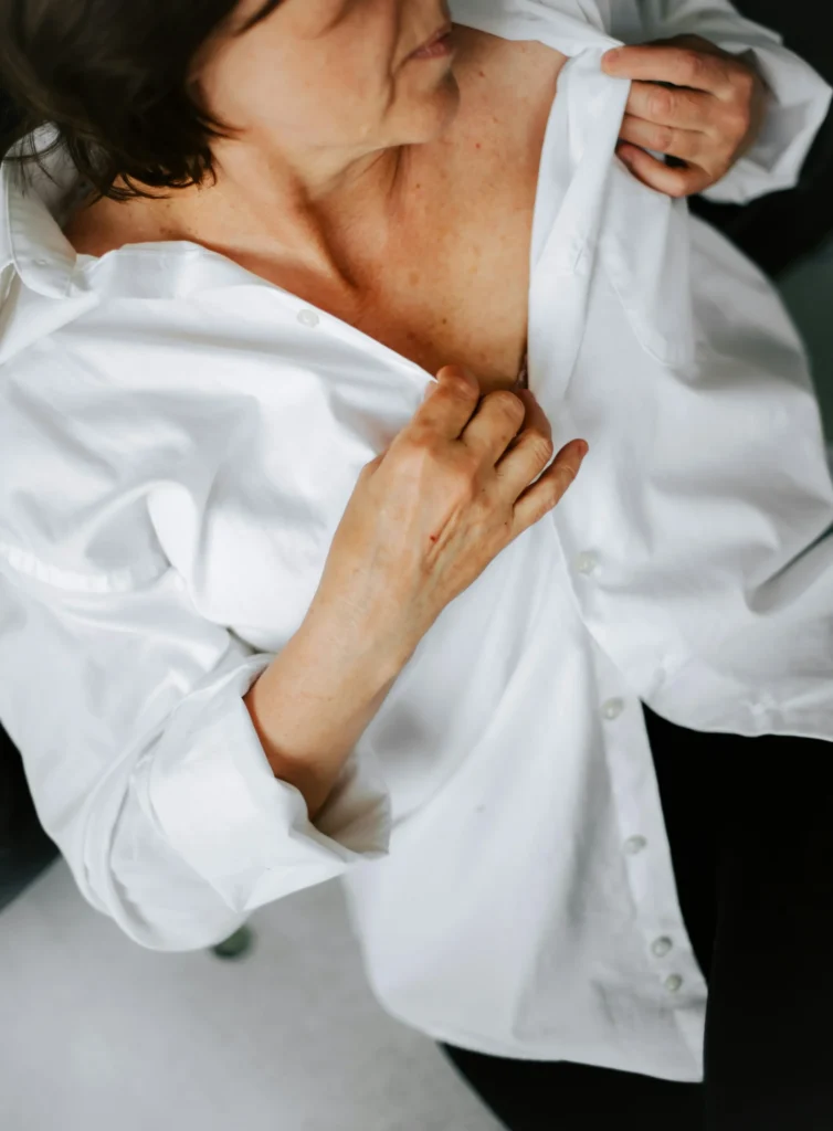 A woman with short brown hair adjusts her white button-up shirt, revealing her neck and chest, against a light background.