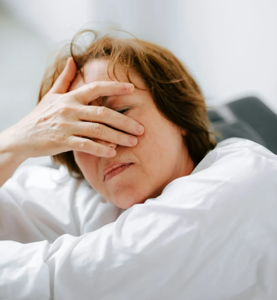 A woman with brown hair covers her eyes with her hand, wearing a white shirt, against a light background, possibly indicating stress or fatigue.