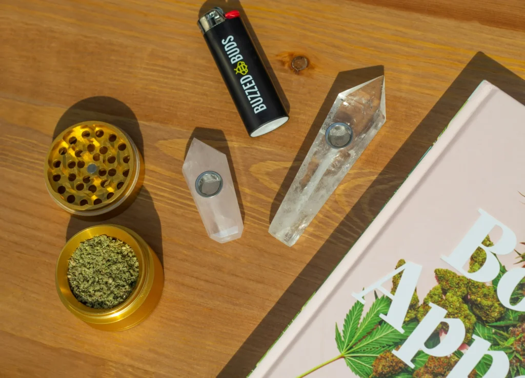 A wooden surface displays a gold grinder with ground cannabis, a "Buzzed Buds" lighter, crystal pipes, and a cannabis-themed book.