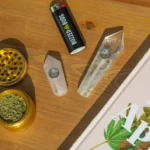 A wooden surface displays a gold grinder with ground cannabis, a "Buzzed Buds" lighter, crystal pipes, and a cannabis-themed book.