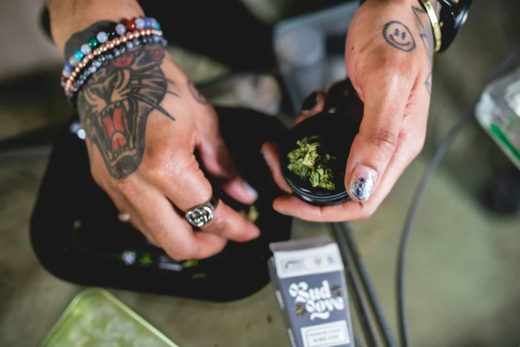 Tattooed hands hold cannabis buds over a black container, with rolling papers and accessories nearby, highlighting cannabis preparation.
