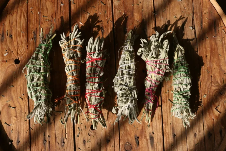 Six sage smudge sticks wrapped with colorful threads (green, orange, red, pink, and white) on a wooden surface, casting shadows.