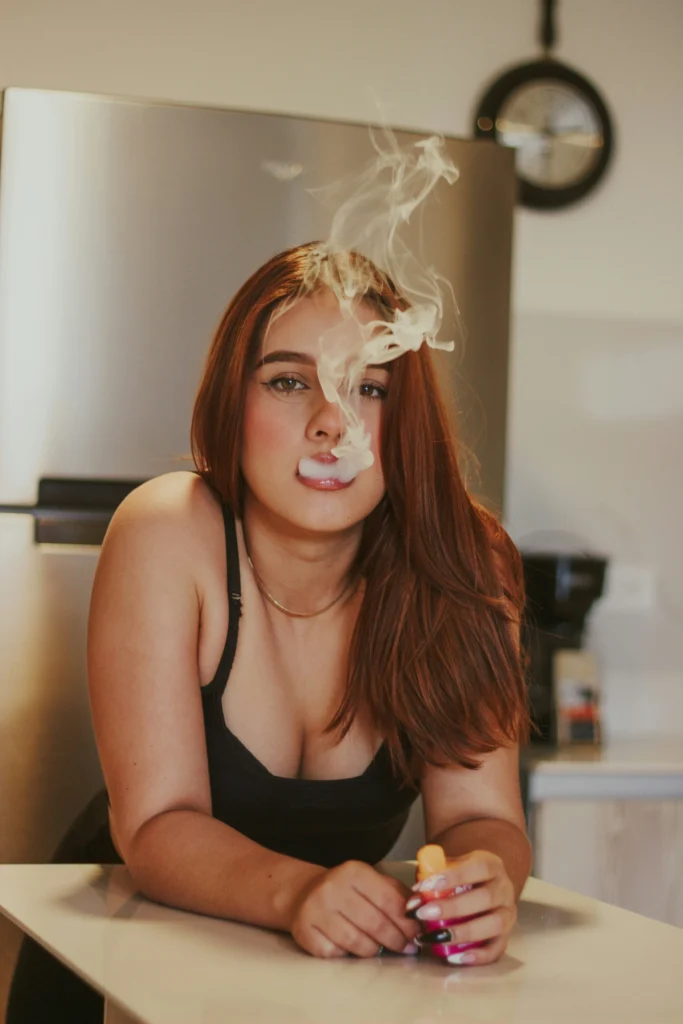 A person with long brown hair vaping in a kitchen, holding a vape pen with vapor in the air.