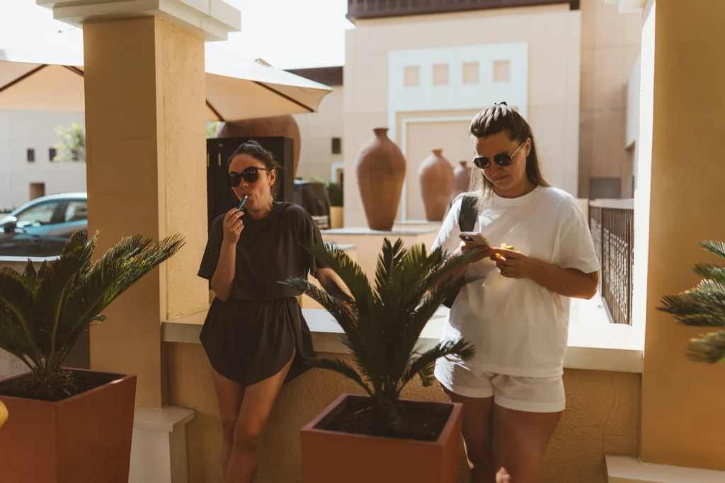 Two people on a sunny patio, one vaping and the other eating a cannabis edible, surrounded by plants.
