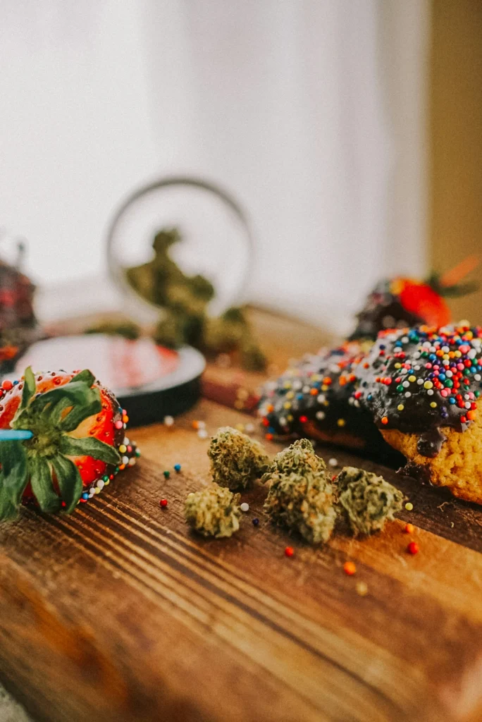 Chocolate-dipped strawberries with sprinkles and a donut alongside cannabis buds on a wooden board.